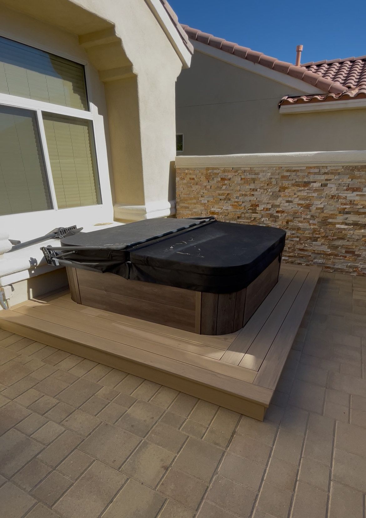 Backyard patio with a covered hot tub on a wooden platform beside a house wall.
