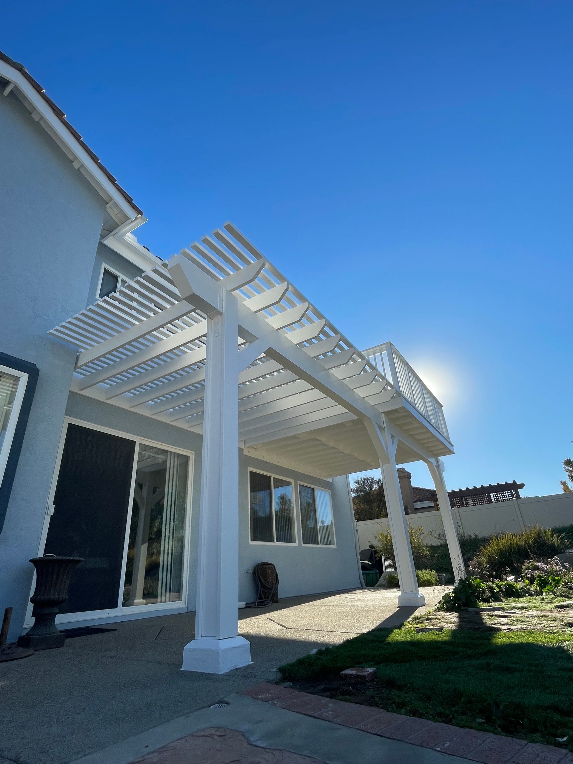 White pergola over a patio beside a house, with blue sky and sunlight.