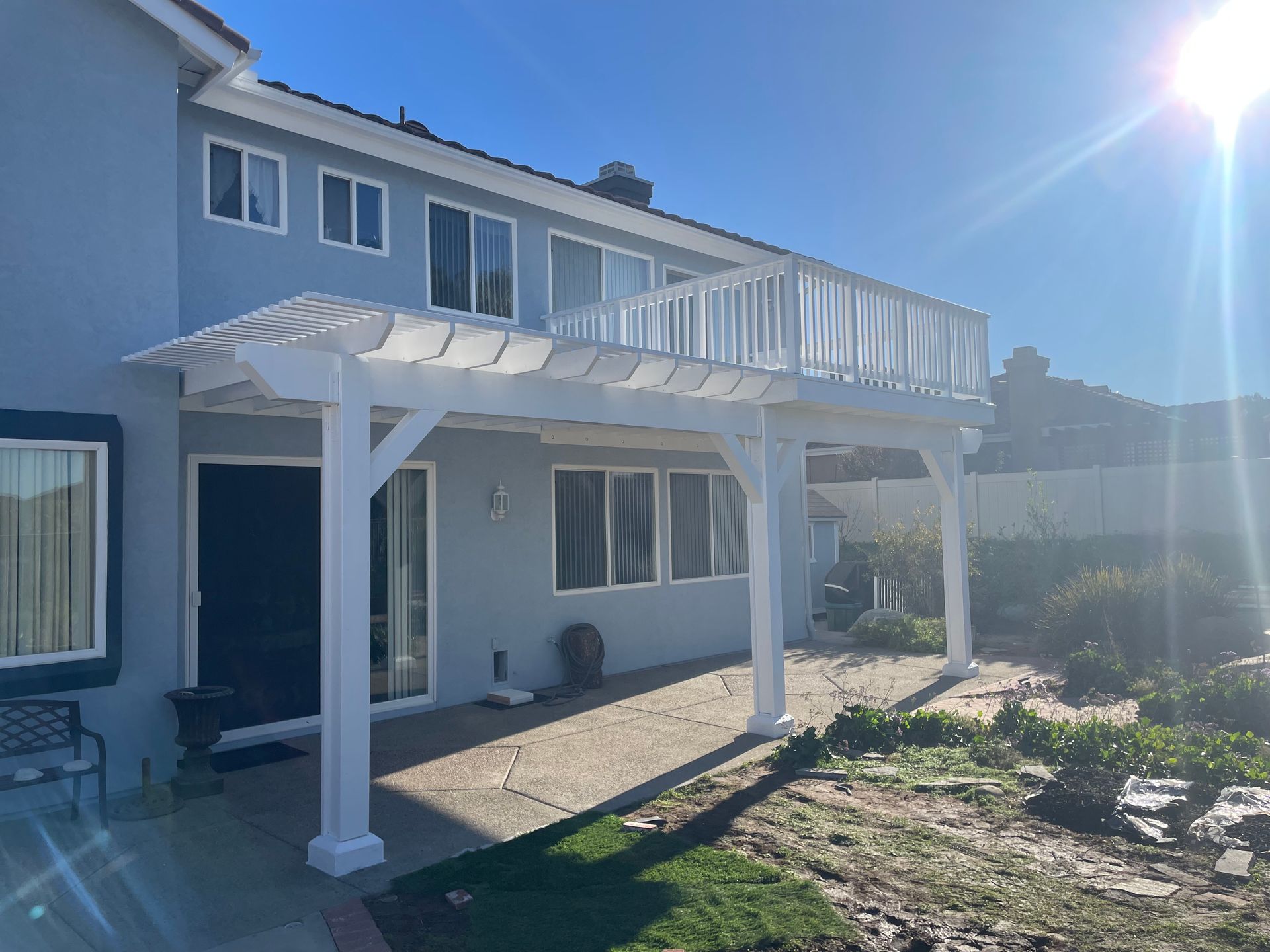 Two-story white house with a balcony and pergola-covered patio in bright sunlight