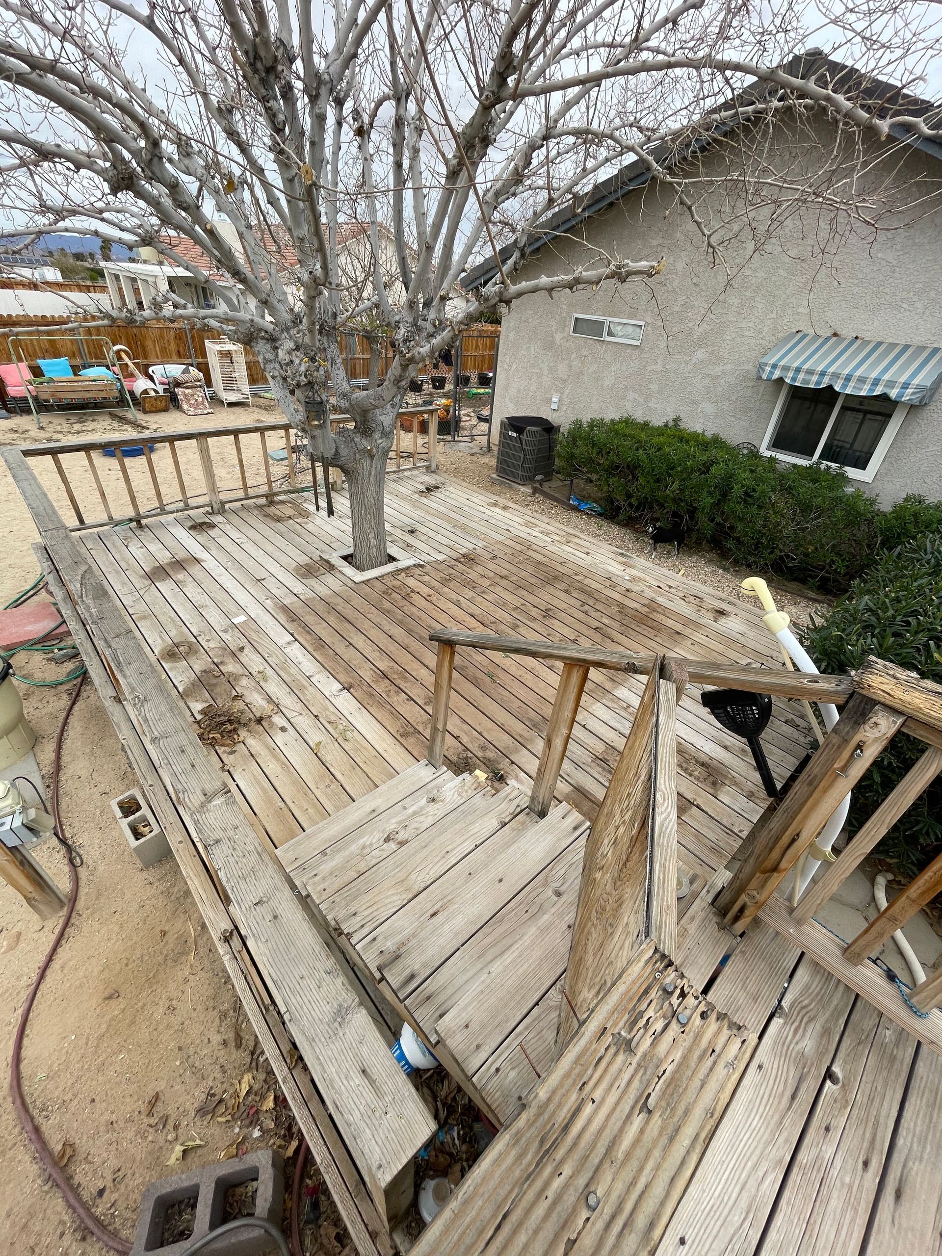 Weathered wooden deck with railings around a leafless tree beside a beige house and small bushes.