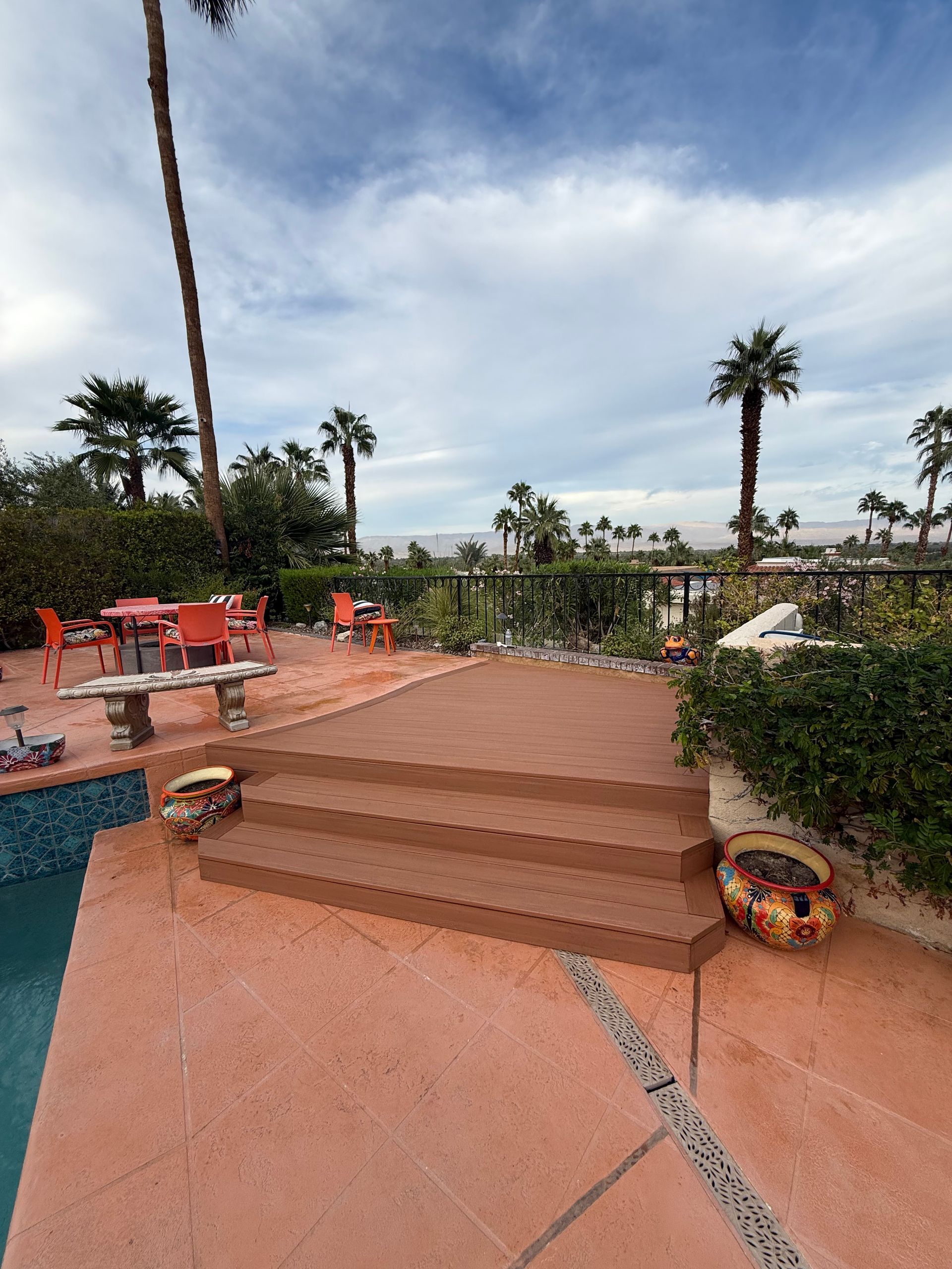 Poolside patio with terracotta steps, red chairs, potted plants, and palm trees overlooking a cloudy sky