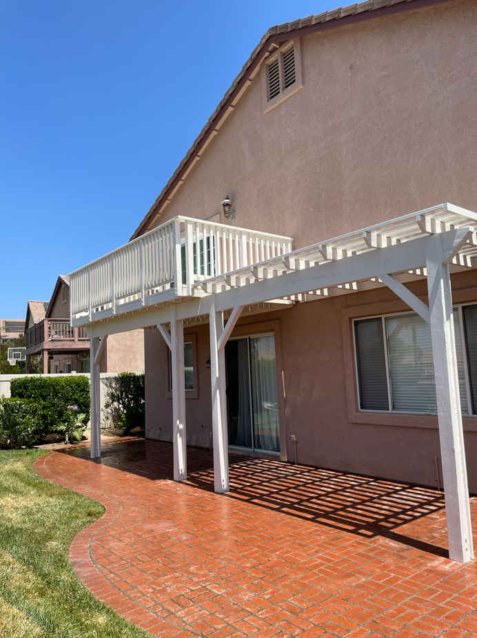 Backyard patio with a brick floor, pergola, and balcony, next to a two-story beige house under a blue sky.