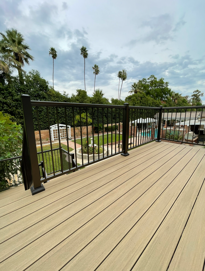 Wooden deck with black railings overlooking a backyard with a pond, trees, and cloudy sky.
