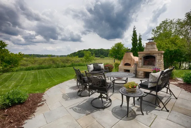 Outdoor patio with stone pizza oven, table, chairs, and field under cloudy sky.
