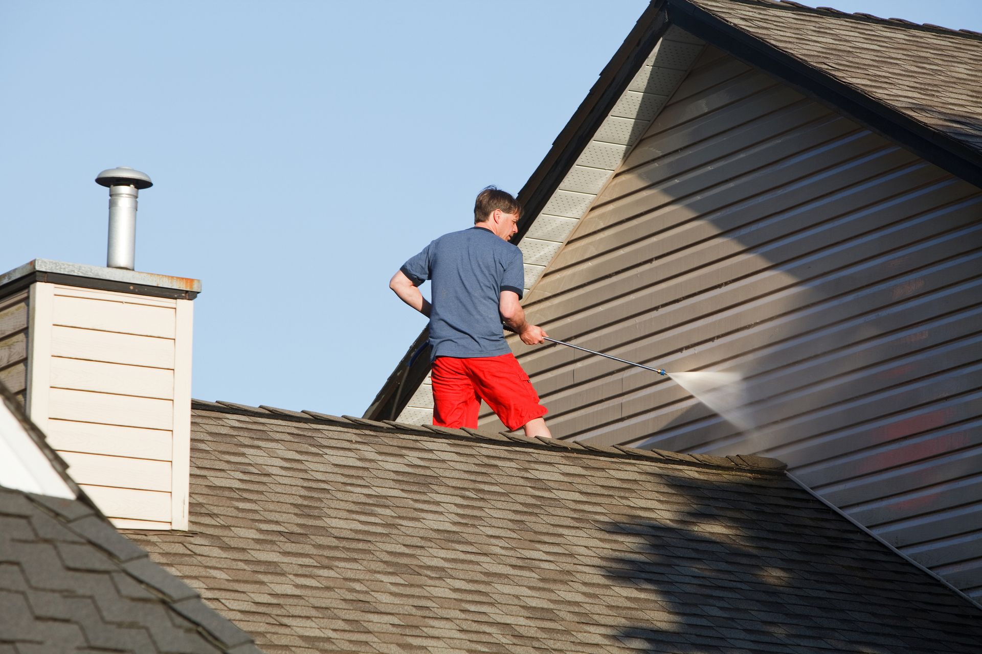 A man is standing on the roof of a house using a pressure washer.