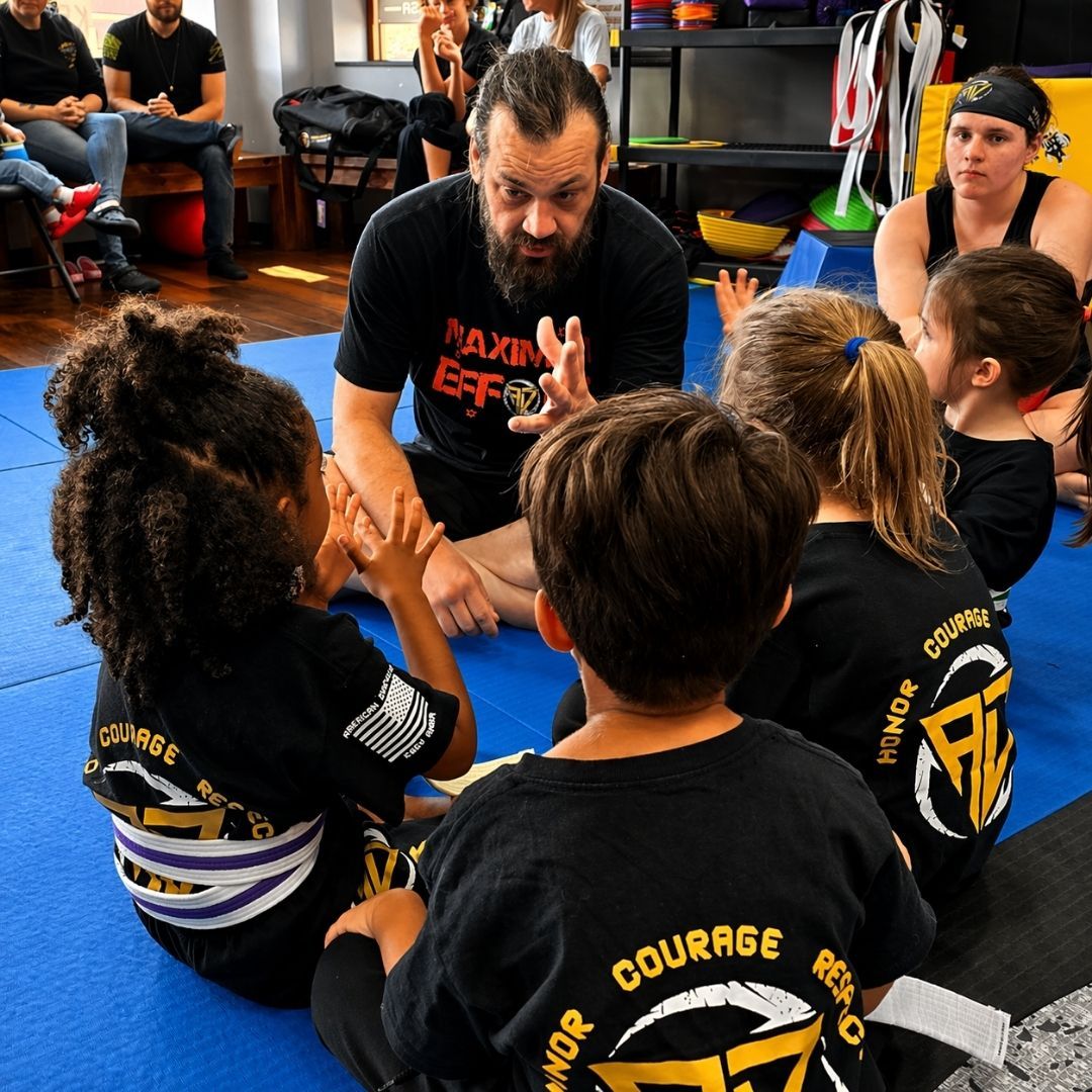 Two young children in blue and black martial arts uniforms grappling on a gym floor, one smiling.