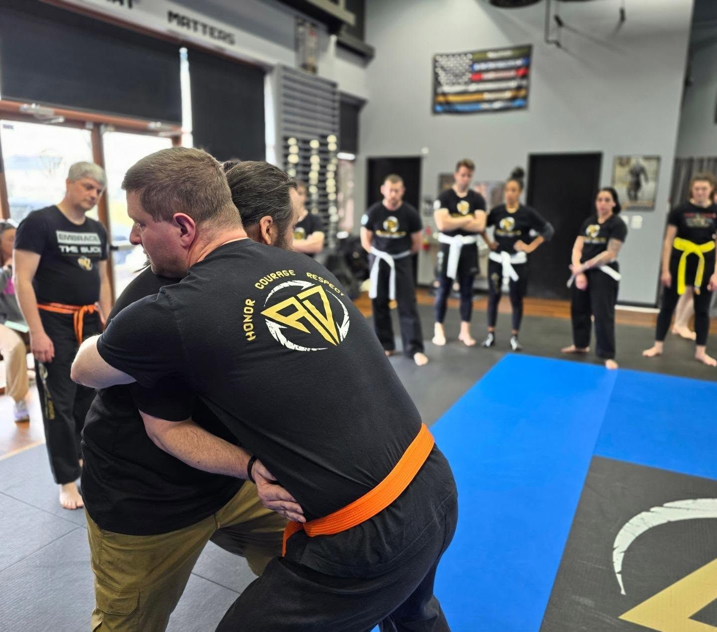 Two men in grappling position on a mat. One embraces the other from behind, both smiling. Gym setting.