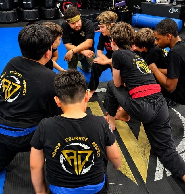 Family in Jiu-Jitsu gis poses. Indoors, blue mats, a smiling woman in the middle, around a seated man with a beard.