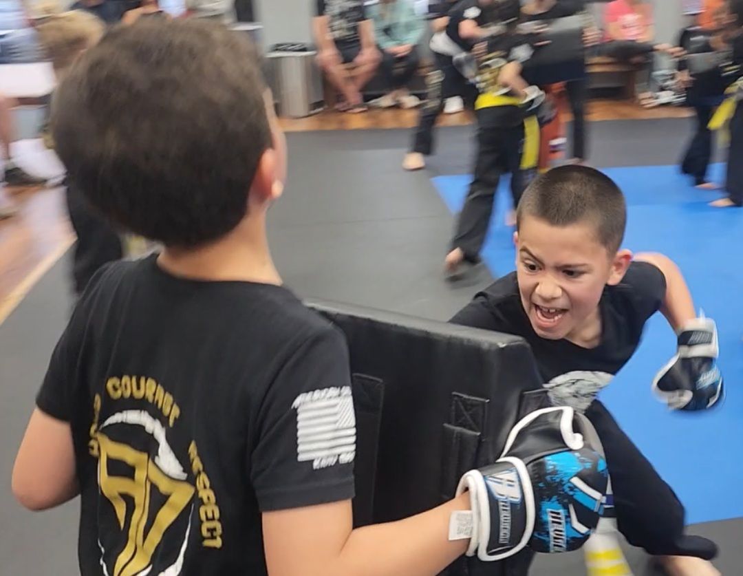 Two children practicing Brazilian Jiu-Jitsu on a mat. One child in white gi has the other in a leg lock. A girl watches.