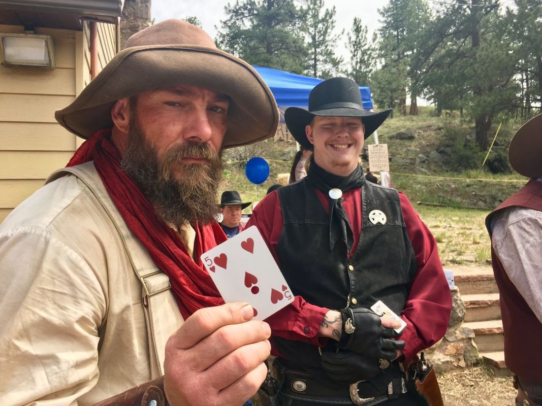 Two men in cowboy attire, one holding playing cards. Outdoor setting with trees and a blue tent.