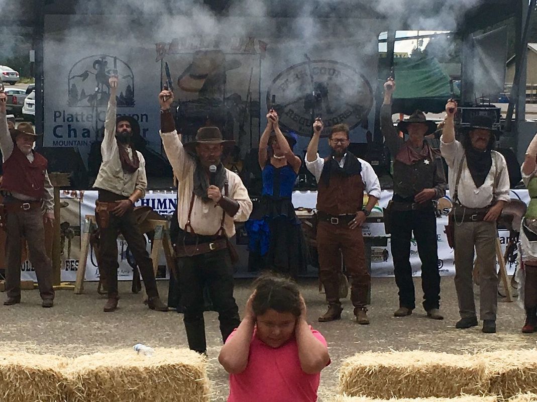 People in Western attire celebrating with guns, smoke, and a child covering ears.