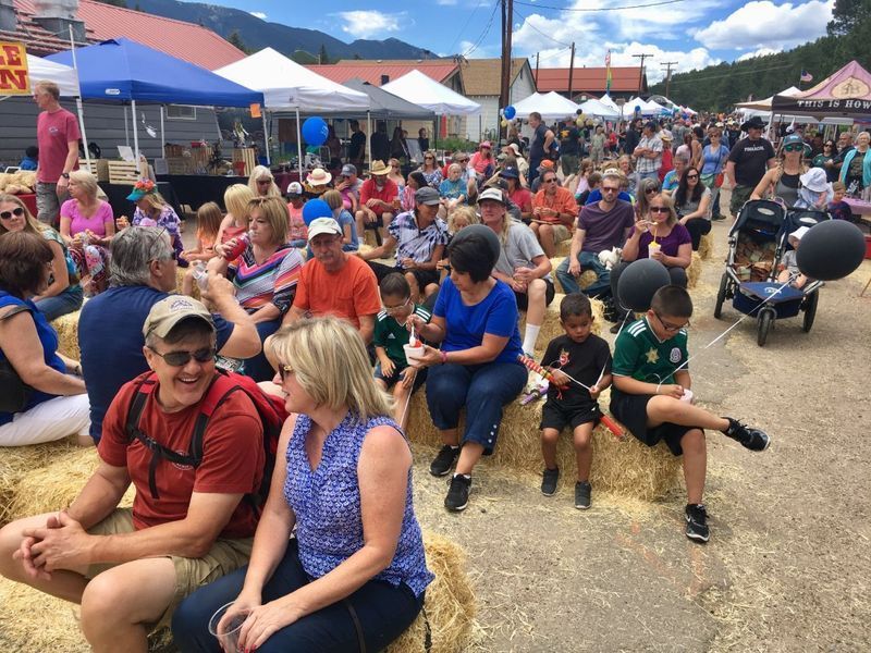 Crowd at an outdoor festival seated on hay bales. People of all ages watch the event; vendors in the background.