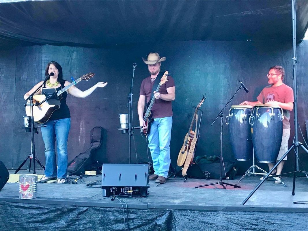 Band on stage performing: woman singing and playing guitar, man playing bass, another on drums. Stage is lit.