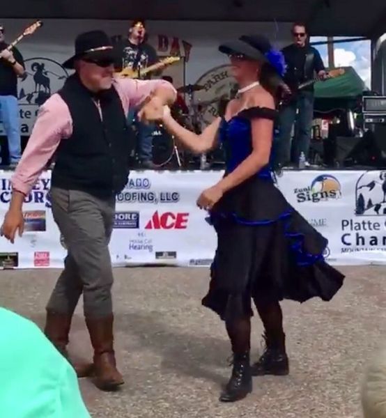 Couple dances on stage with a band. Man in cowboy hat, vest, woman in blue dress, black skirt.