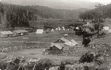 Historical black and white photo of a small settlement in a valley, with wooden buildings, tents, and a fence.
