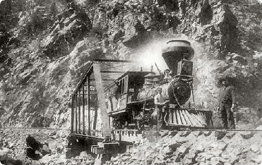 Steam engine train on tracks carved into a mountain side, with a man standing nearby.