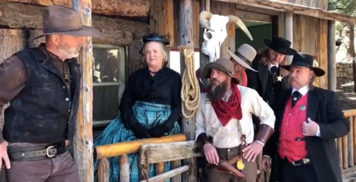 People in Western attire pose on a wooden porch, cow skull above.