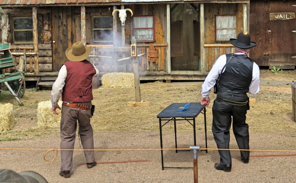 Two people in cowboy attire shooting guns in front of a western-style building. One aims, the other observes by a table.