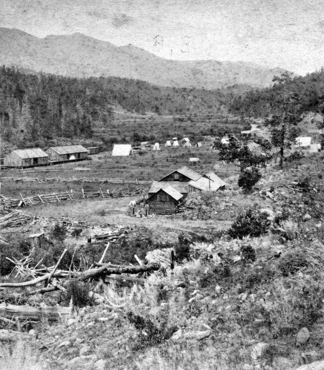 Black and white photo of a small settlement in a valley, with simple buildings and mountains in the background.