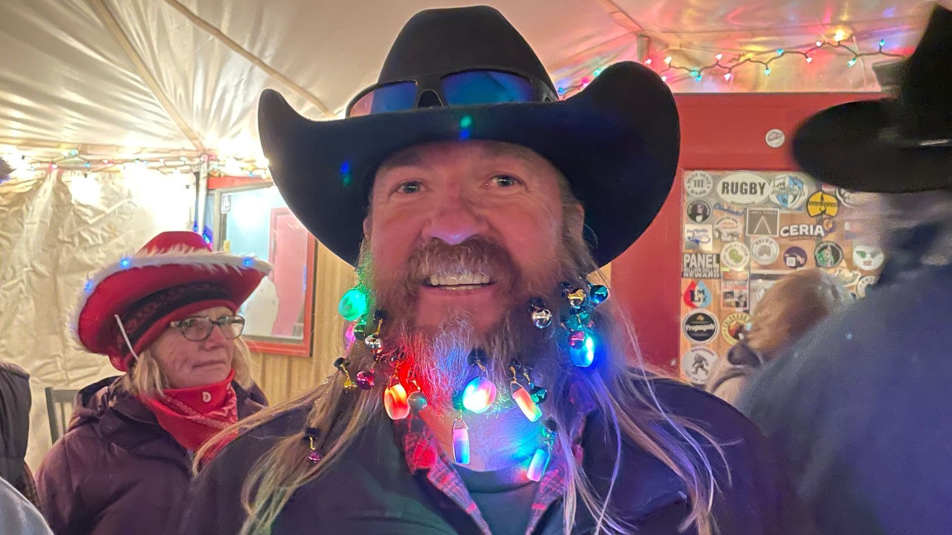 Man in cowboy hat with beard decorated with Christmas lights smiles outdoors.