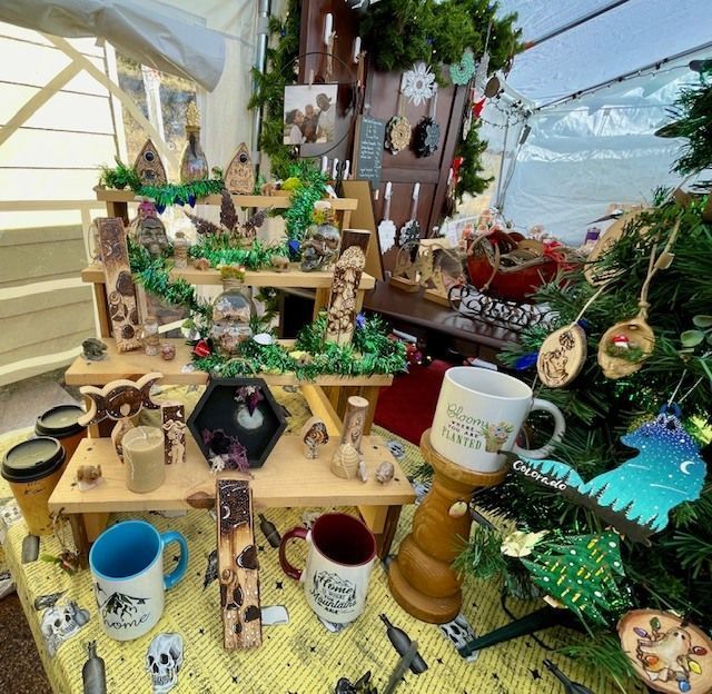 A craft booth with wooden carvings, mugs, and ornaments displayed on tiered shelves and a Christmas tree.