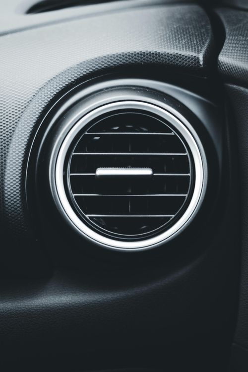 A circular, black automotive air conditioning vent with silver trim, set into a dark, textured dashboard.