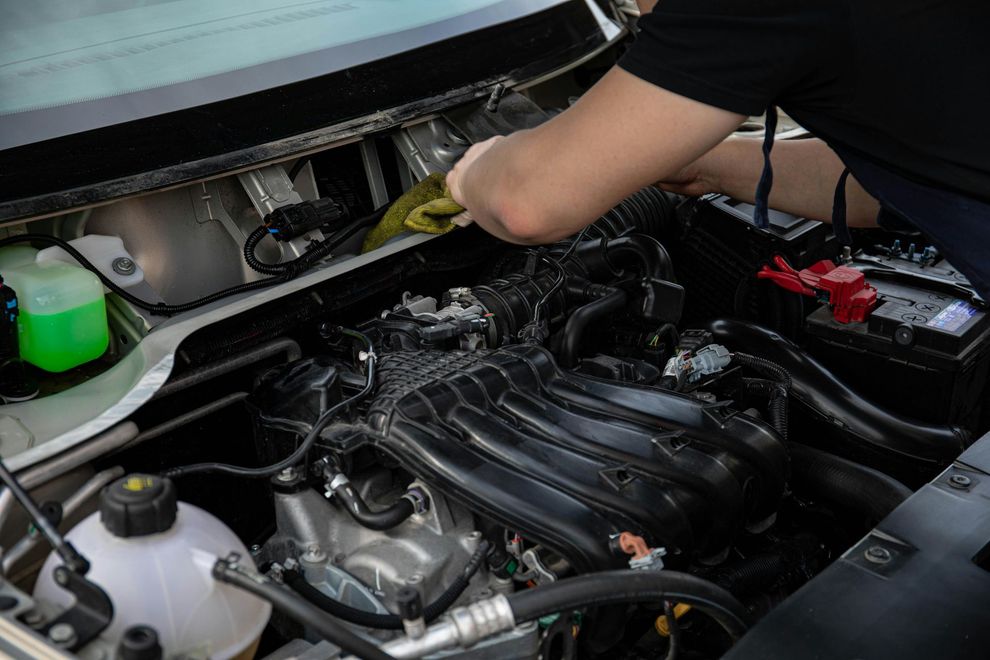A person in a black shirt reaches into the engine bay of a car to perform maintenance or cleaning.