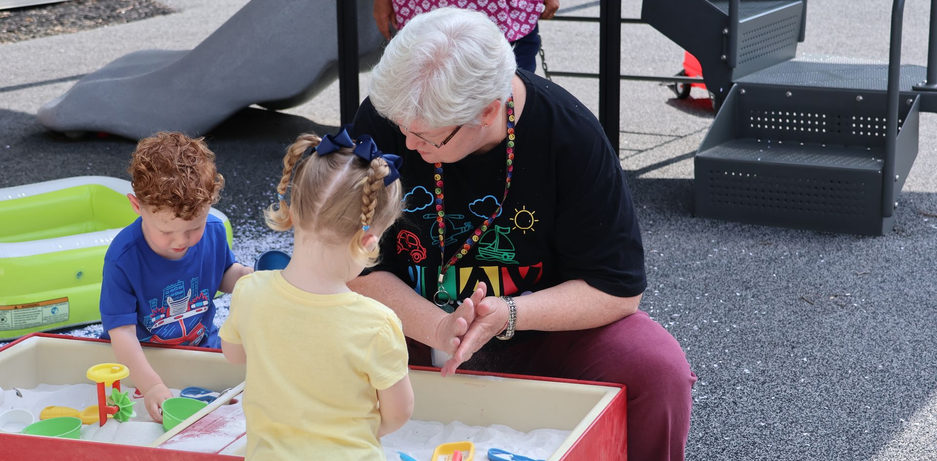 A woman plays in a sensory box with two young children on a playground.