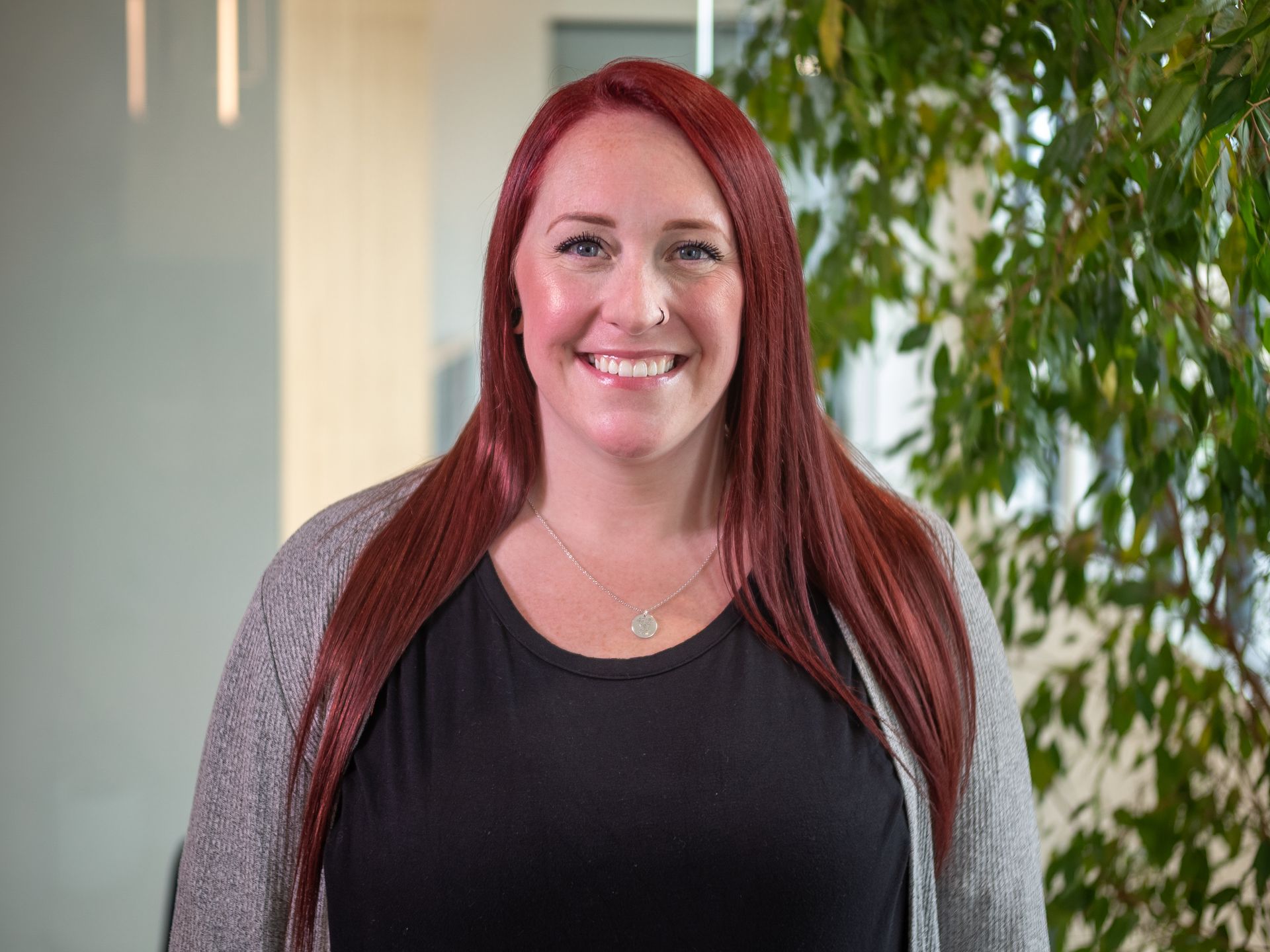 Smiling person with long red hair in a black top and gray cardigan, standing indoors near plants.