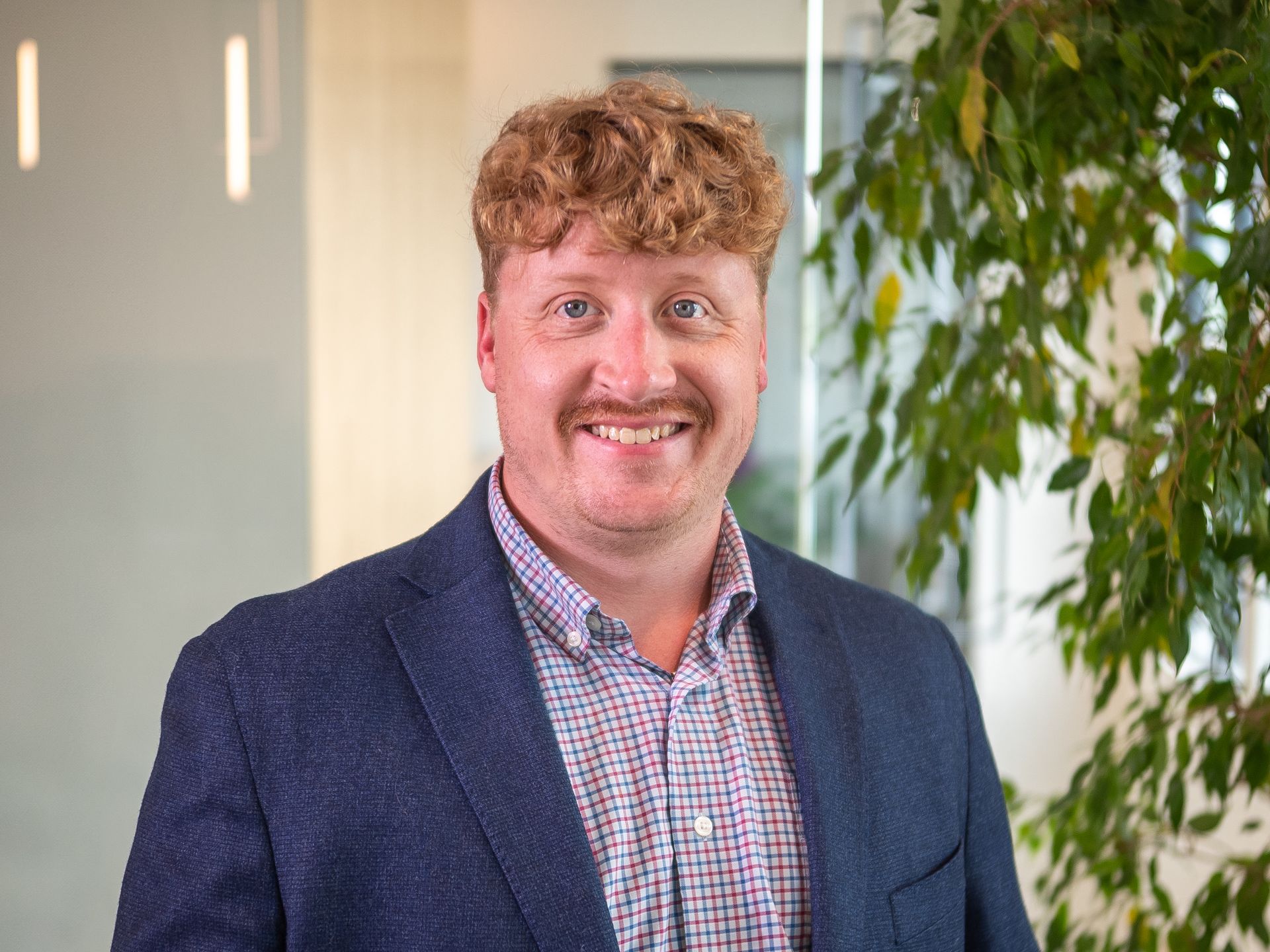 Smiling man in a blue blazer and checkered shirt standing indoors beside a plant.