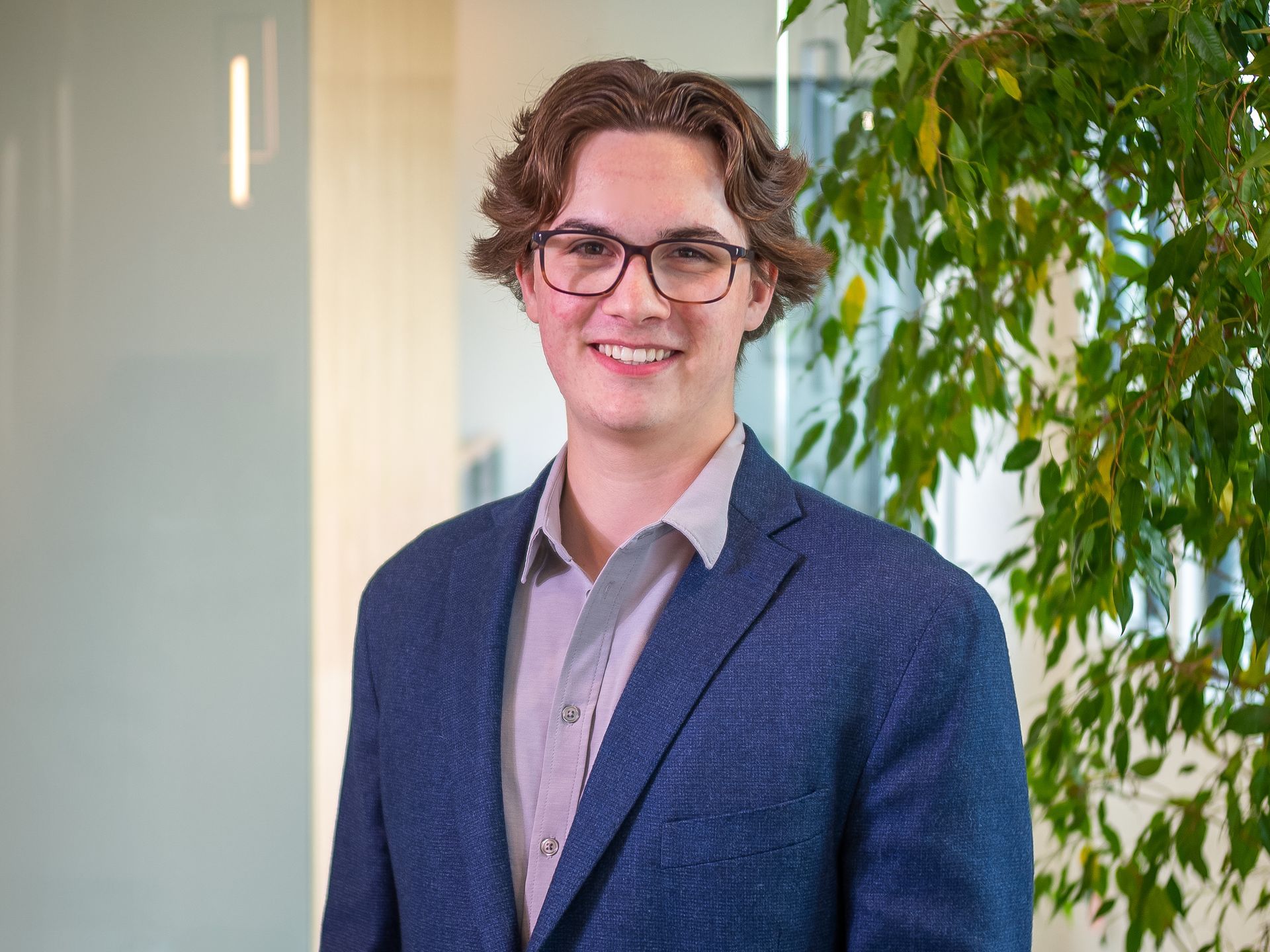 Smiling person in glasses and a blue blazer standing indoors near a leafy plant.