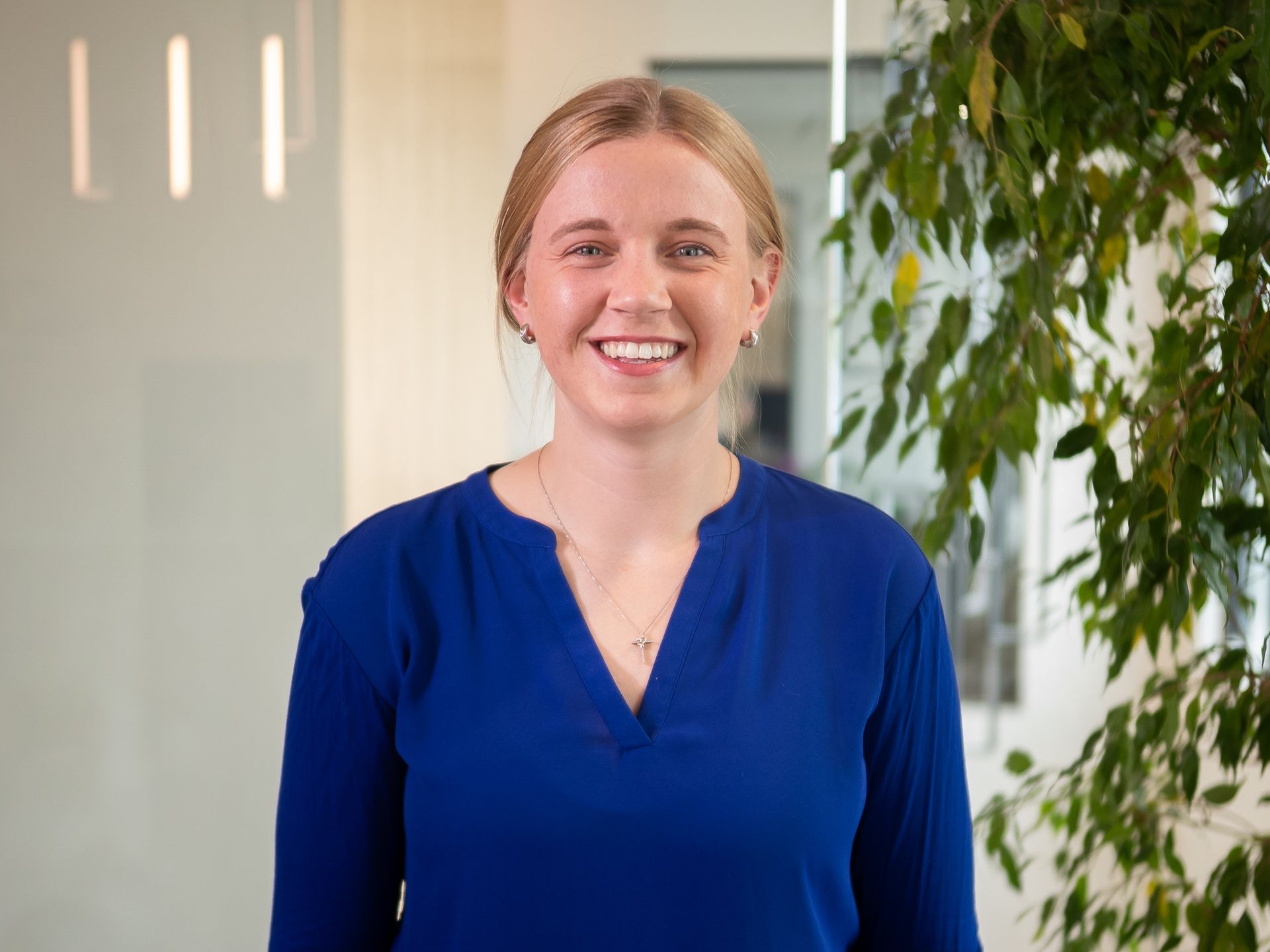 Smiling woman in a blue blouse standing in an office with a plant in the background