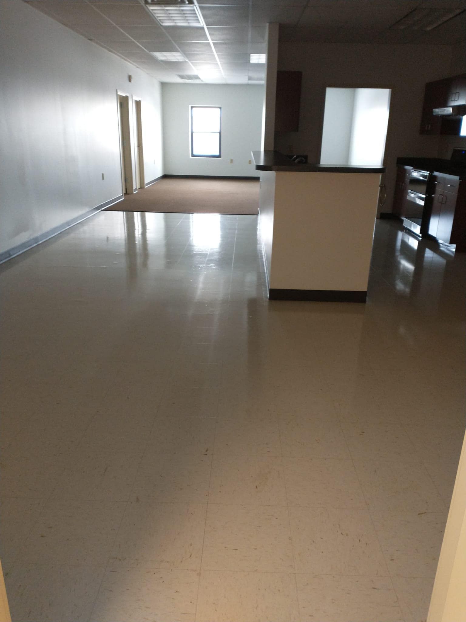 Empty office space with white walls, tan floors, and a small kitchen area.