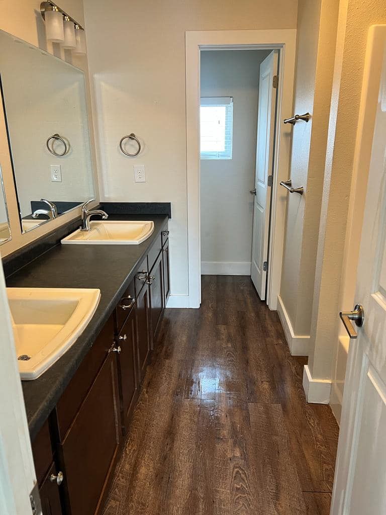 Bathroom with dark wood cabinets, dual sinks, and a long hallway leading to a window and door.