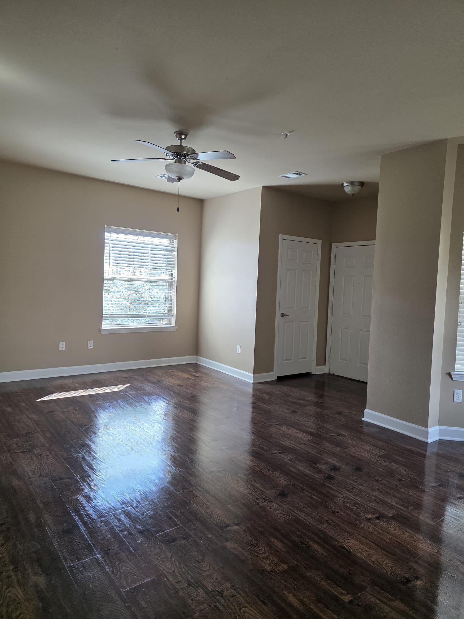 Empty living room with dark wood floors, beige walls, two white doors, and a ceiling fan.