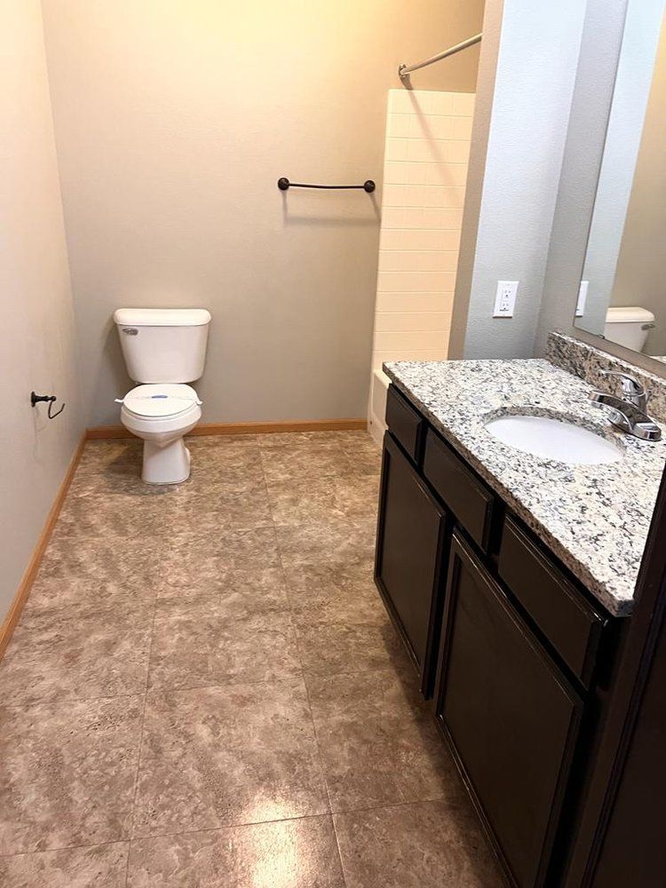 Bathroom with toilet, vanity, and shower. Tan walls, brown cabinets, and patterned flooring.