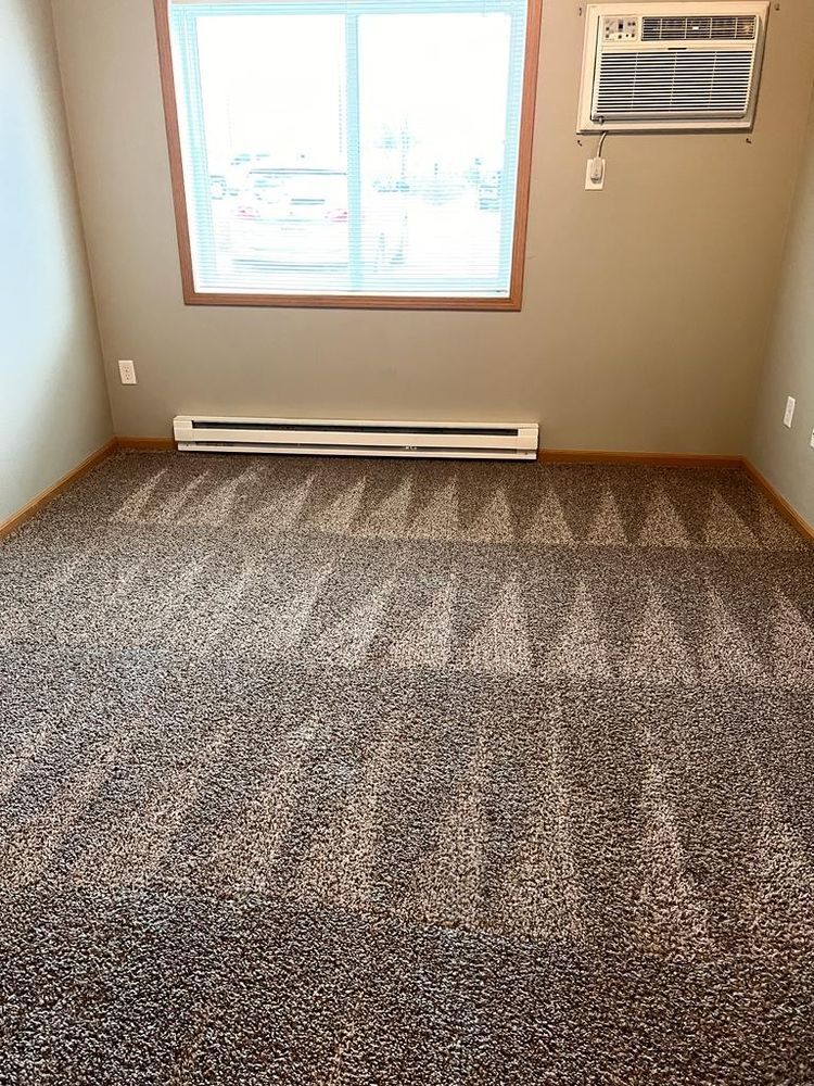 Room with patterned carpet, window, baseboard heater, and wall-mounted air conditioner.