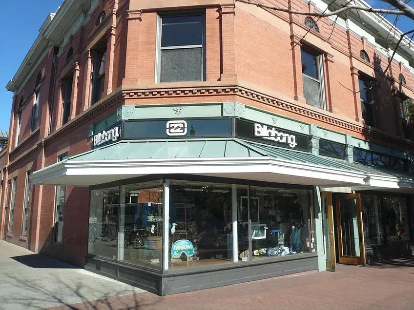 Street corner storefront in a brick building with large windows and green trim, sunlit sidewalk outside.