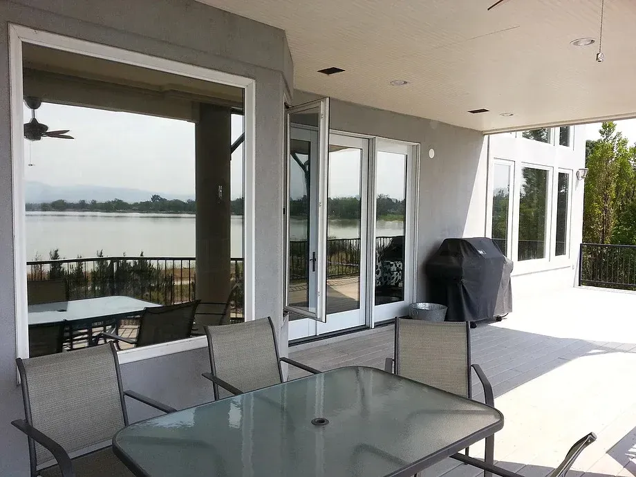 Patio with glass table and chairs beside a lake, with sliding doors and a covered grill on a bright terrace