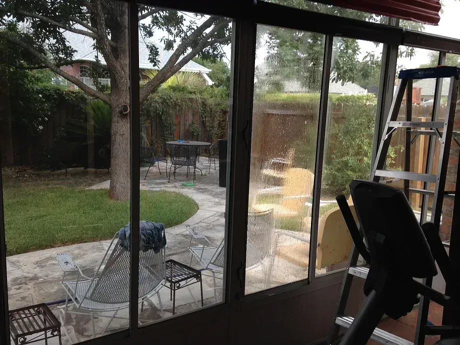 Backyard patio seen through sliding glass doors, with chairs, table, grill, and yard beyond
