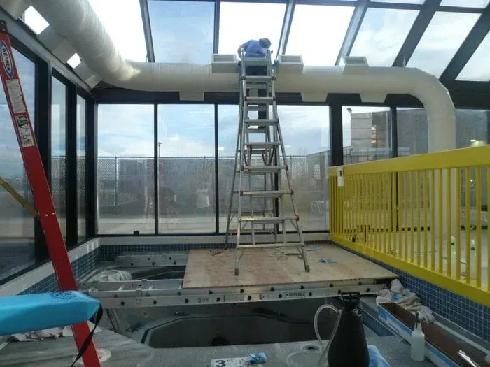 Ladder in a glass-walled indoor space with a skylight, red beam, yellow railing, and open floor panels