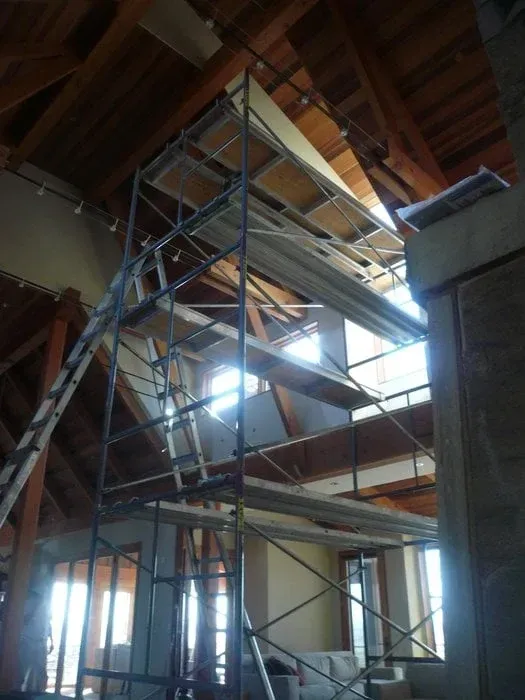 Scaffolding inside an unfinished building with exposed wooden roof beams and bright light through windows