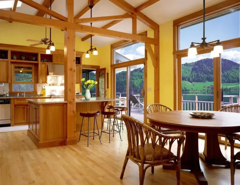 Bright wood cabin interior with kitchen, dining table, and large windows overlooking mountains and lake