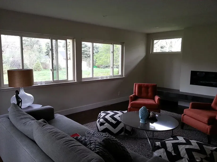 Modern living room with gray sofa, red chairs, round coffee table, chevron ottoman, and large windows