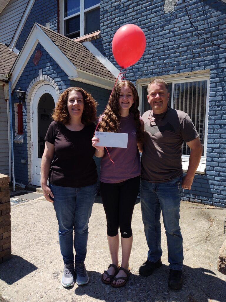 A woman holding a check and a red balloon stands with her parents in front of a blue brick house.