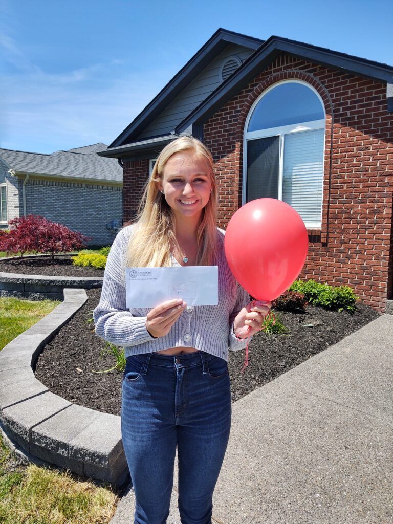 A woman holding a check and a red balloon in front of a brick house.