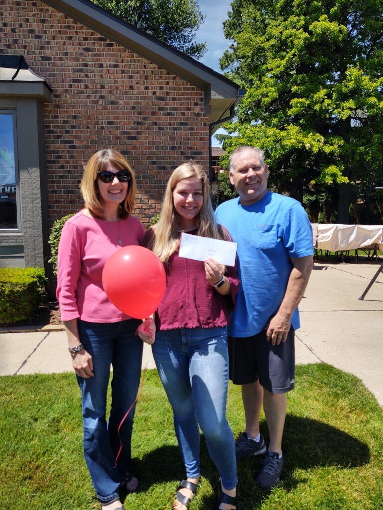 A woman holding a red balloon and a check stands with her parents.