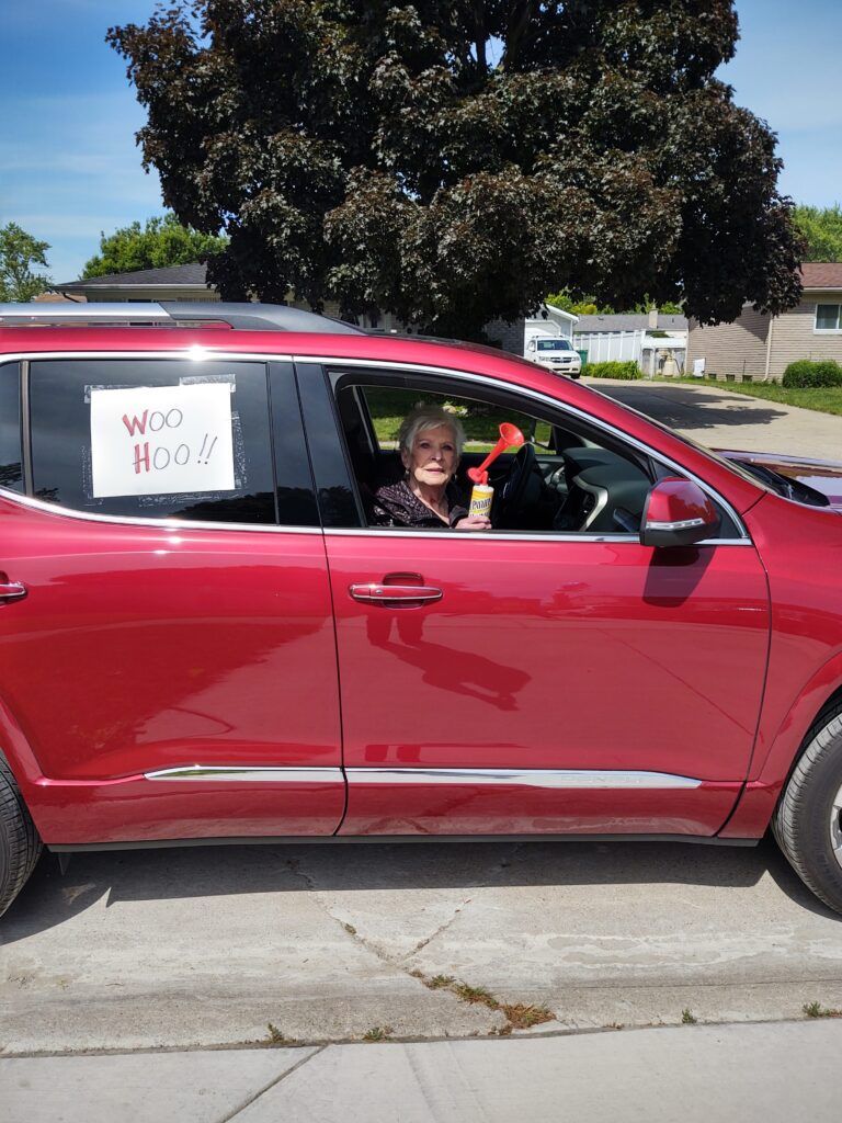 A woman sits in a red car with a sign in the window that says woo hoo.