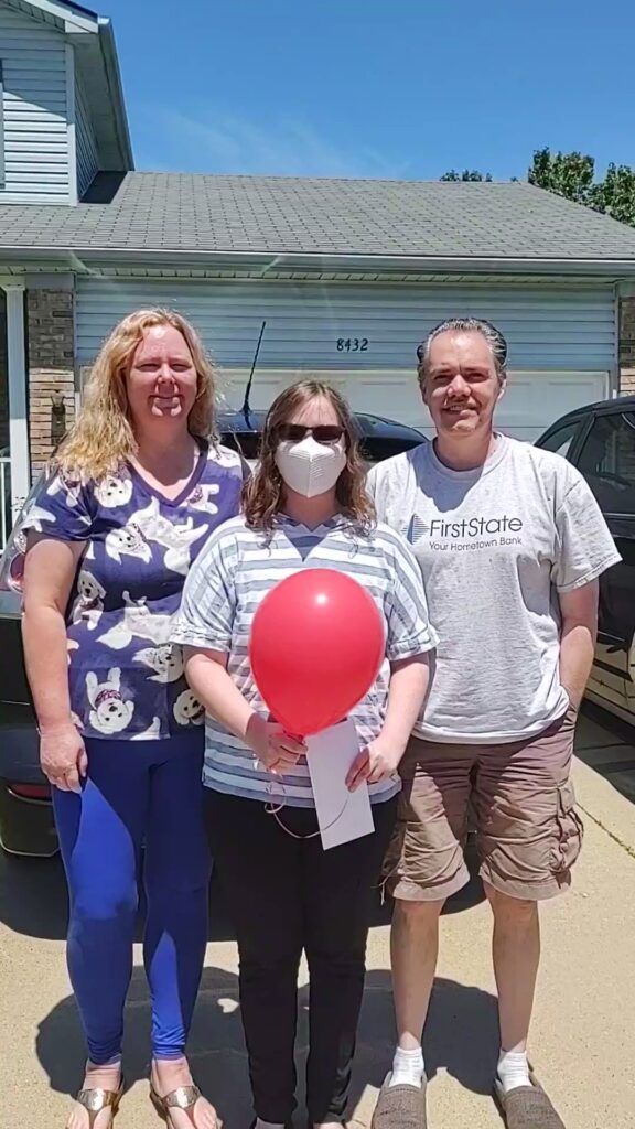 A person holding a check and a red balloon stands next to their parents.