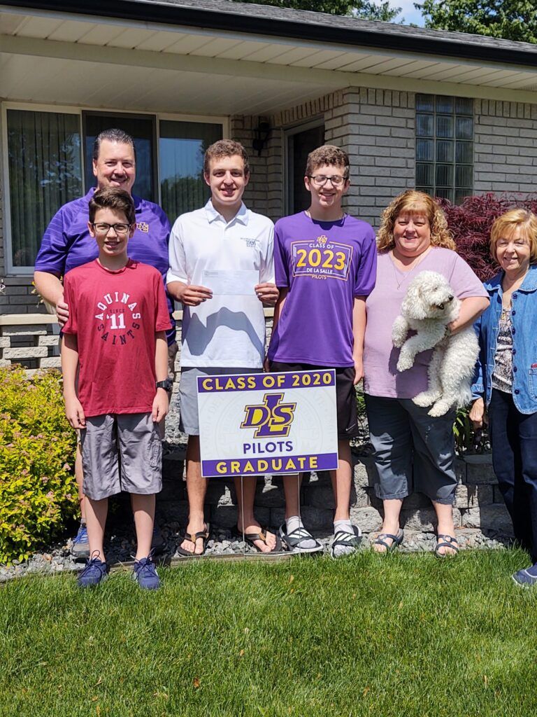 A group of people standing in front of a house holding a sign that says Class of 2020 Pilots Graduate.
